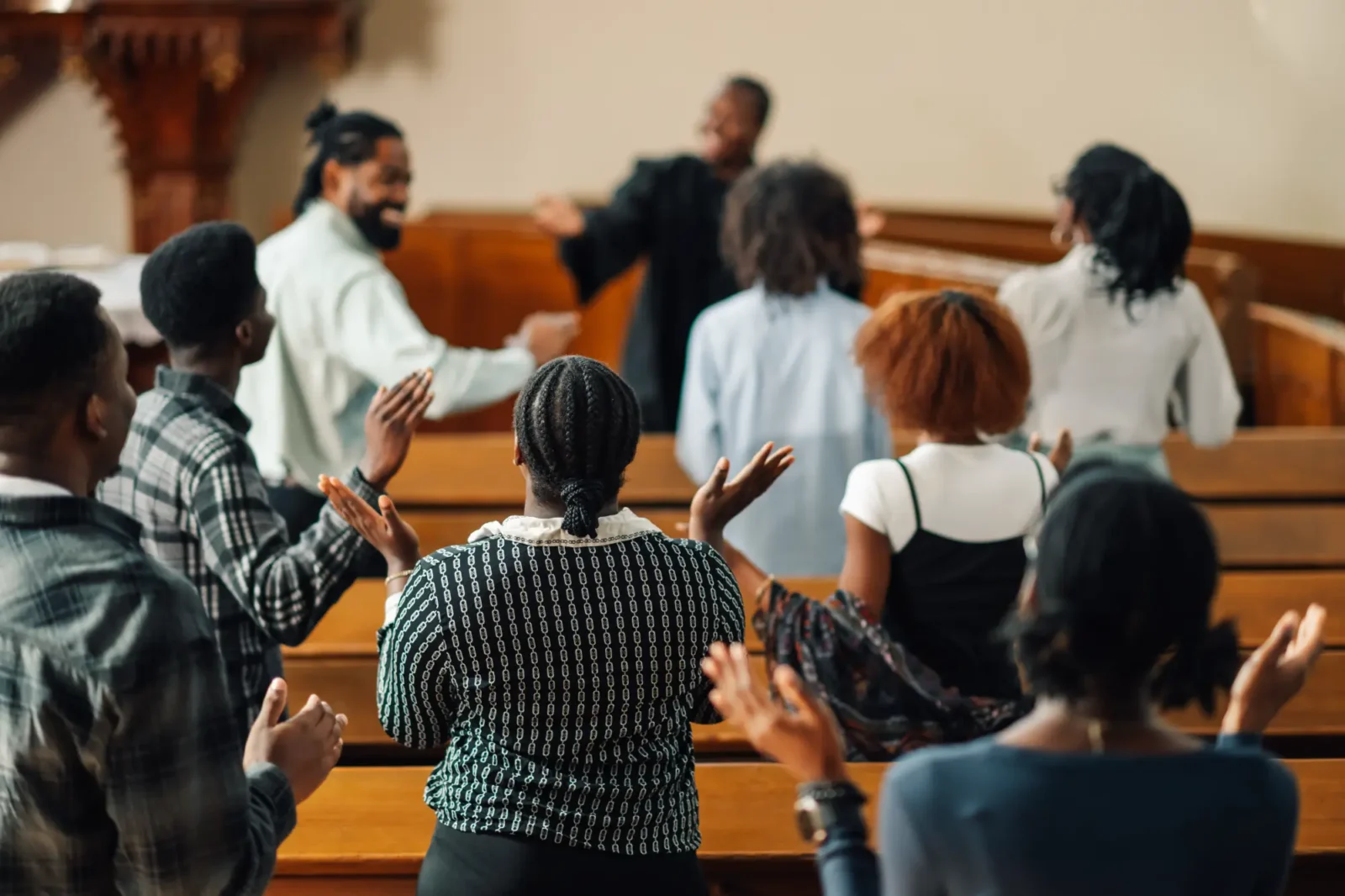 Members of a church congregation sit in wooden pews, enthusiastically clapping and raising their hands toward a speaker in a black robe.