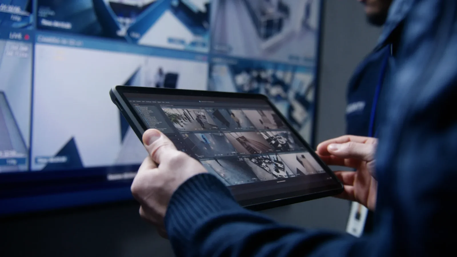 A person in a dark blue sweater holds a tablet displaying a grid of live security camera feeds, with large surveillance monitors visible behind them.