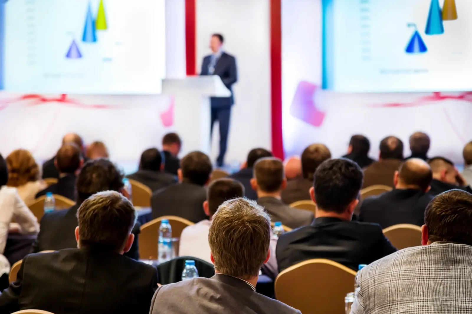 A large audience of professionals in business attire sits, looking toward a speaker presenting at a podium in a conference room.