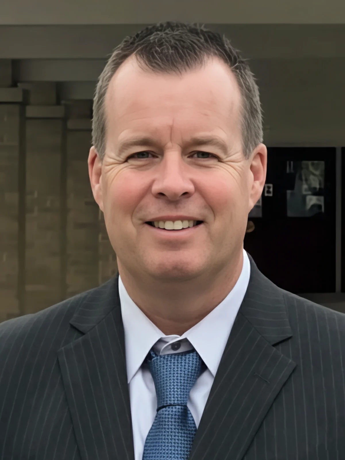 A professional headshot of John Ahern, who is smiling and wearing a dark pinstripe suit and blue patterned tie against a neutral outdoor background.