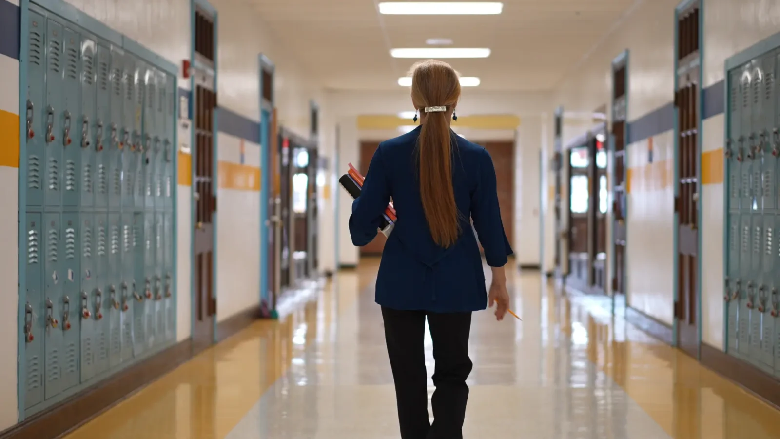 A woman with long hair walks down a school hallway, carrying several books and a pencil in her hand.