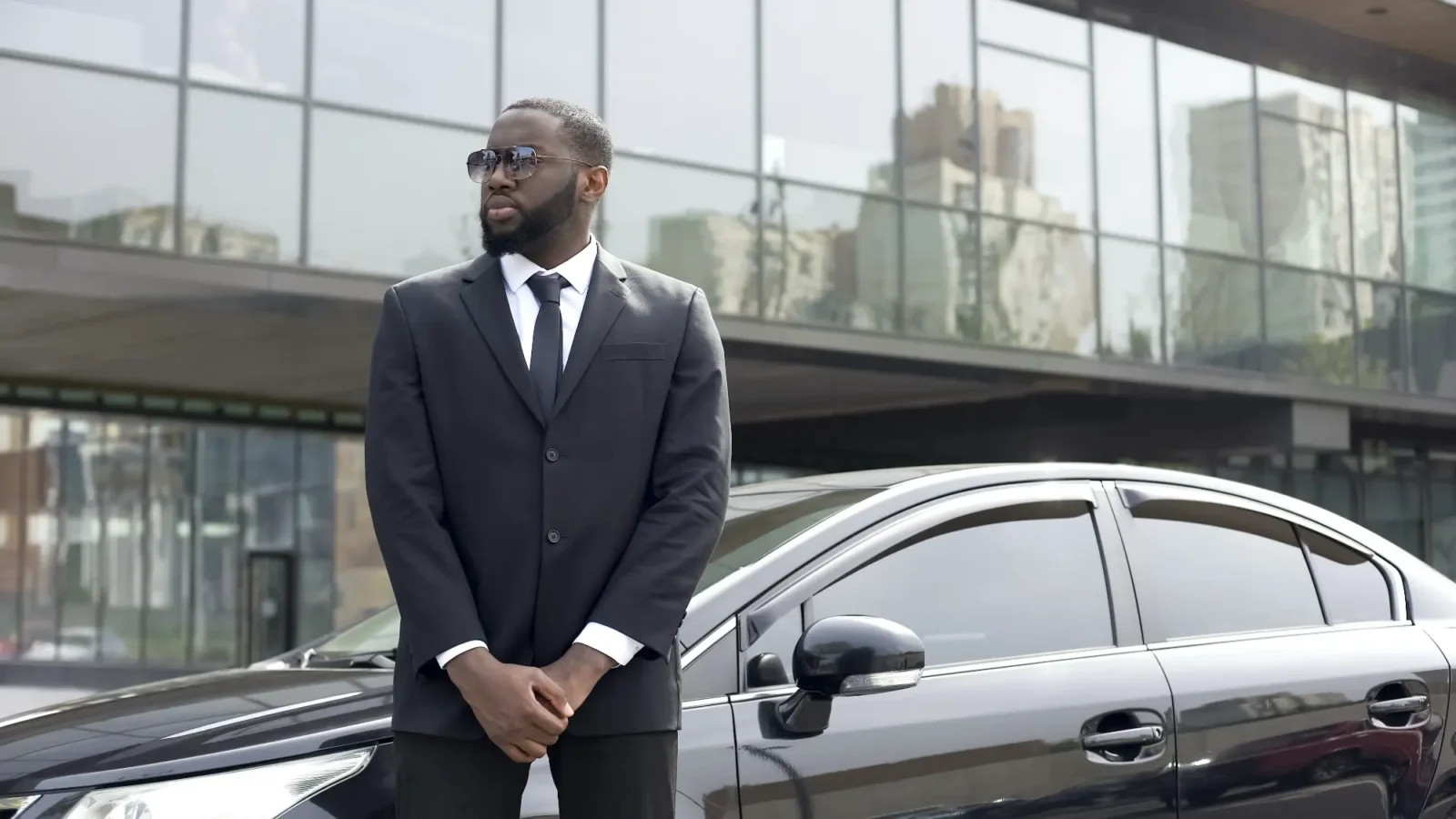 A male security professional in a black suit and sunglasses stands formally next to a black car outside a glass building.