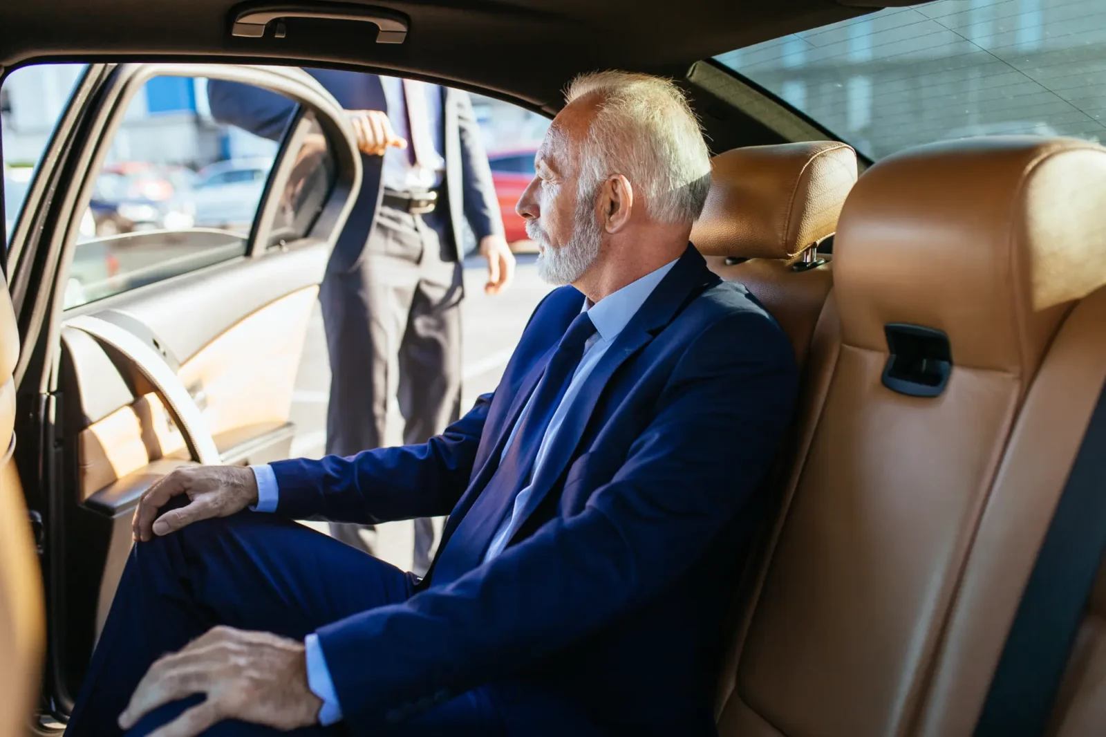 A businessman in a dark suit sits in the back seat of a luxury car, looking out the open door where a suited attendant waits.