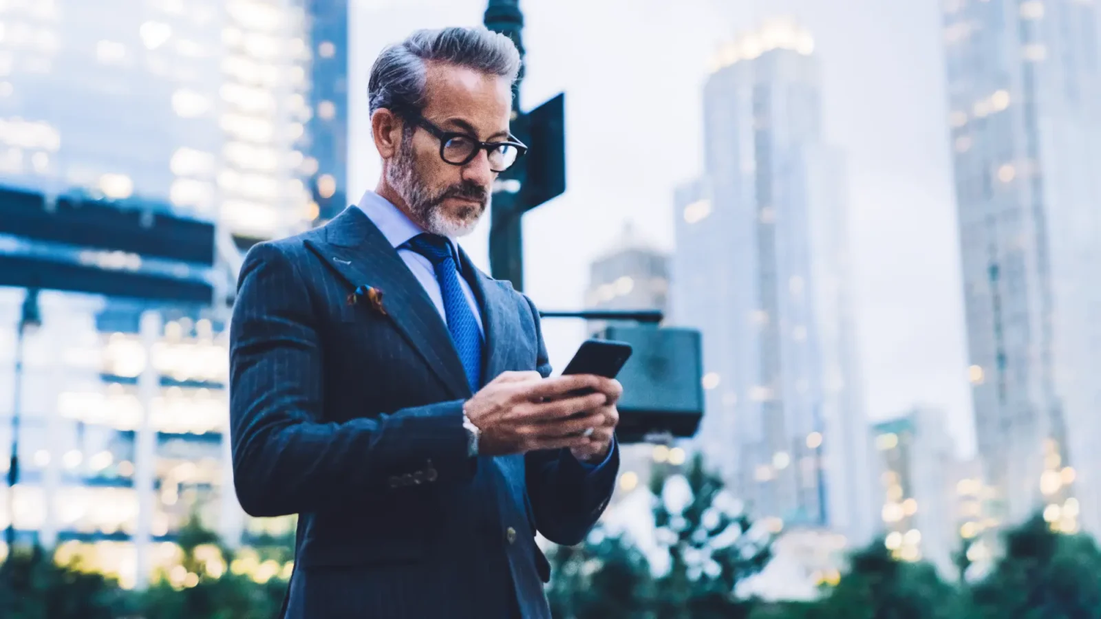 A man in a dark pinstripe suit and glasses stands on a city street, focused on content on his black smartphone.