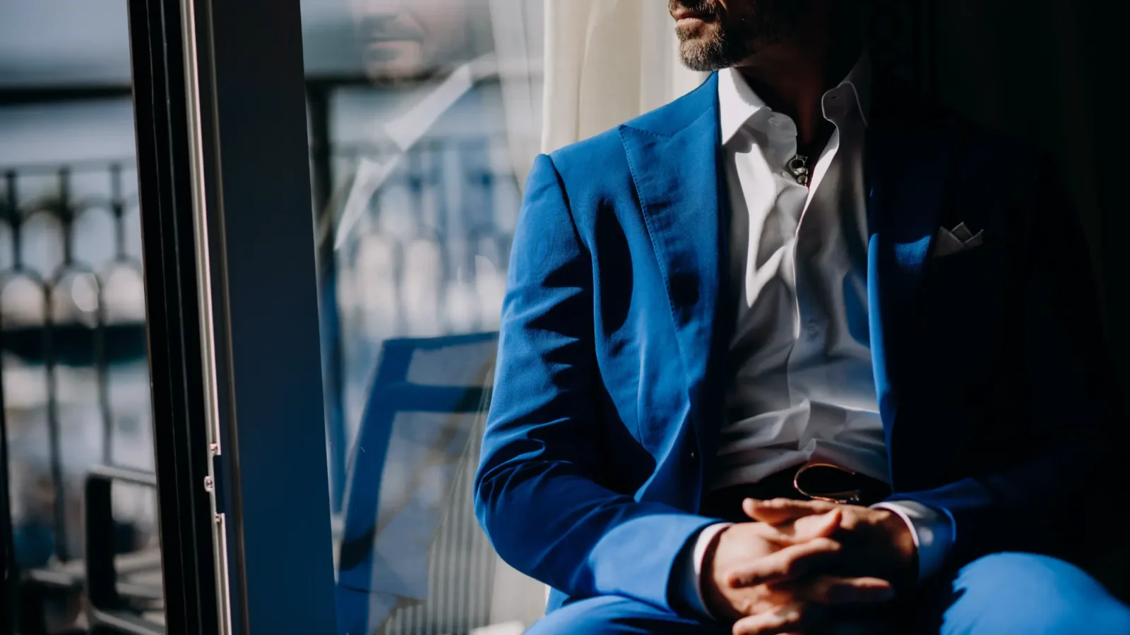 A man in a bright blue suit sits by a window in dramatic light, with his hands clasped and a focused expression.