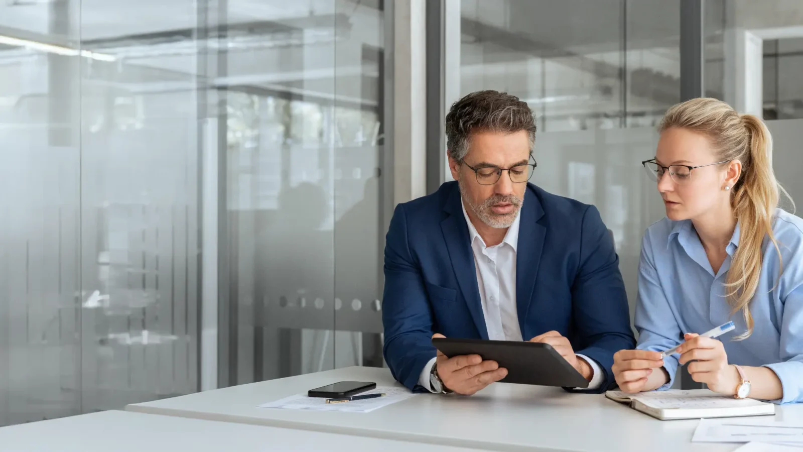 A male business advisor reviews a tablet with a focused female client, who holds a pen and looks at the screen in a modern office.