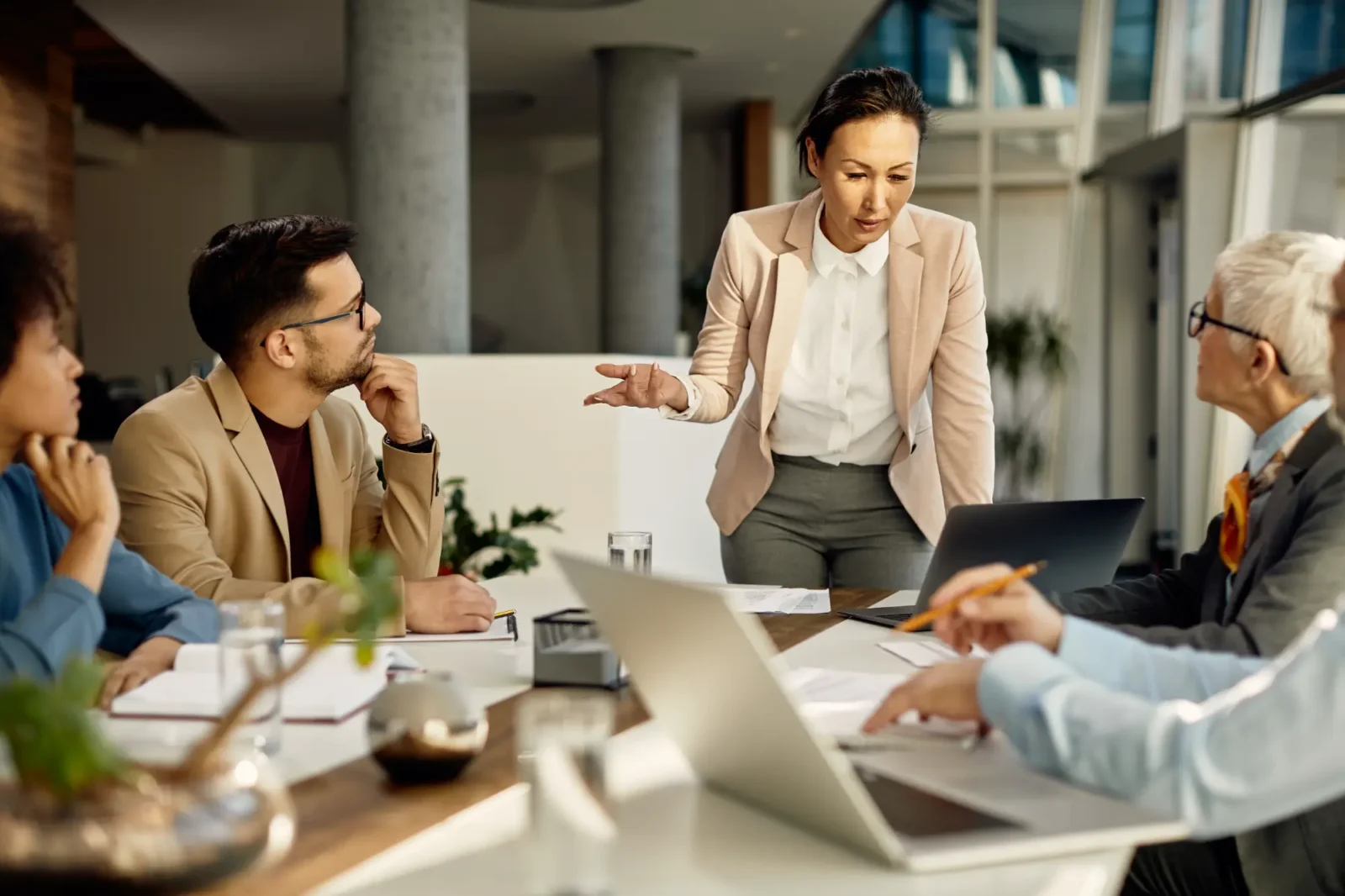 A woman in a business suit stands, leaning over a conference table to speak to four colleagues during a collaborative business meeting.