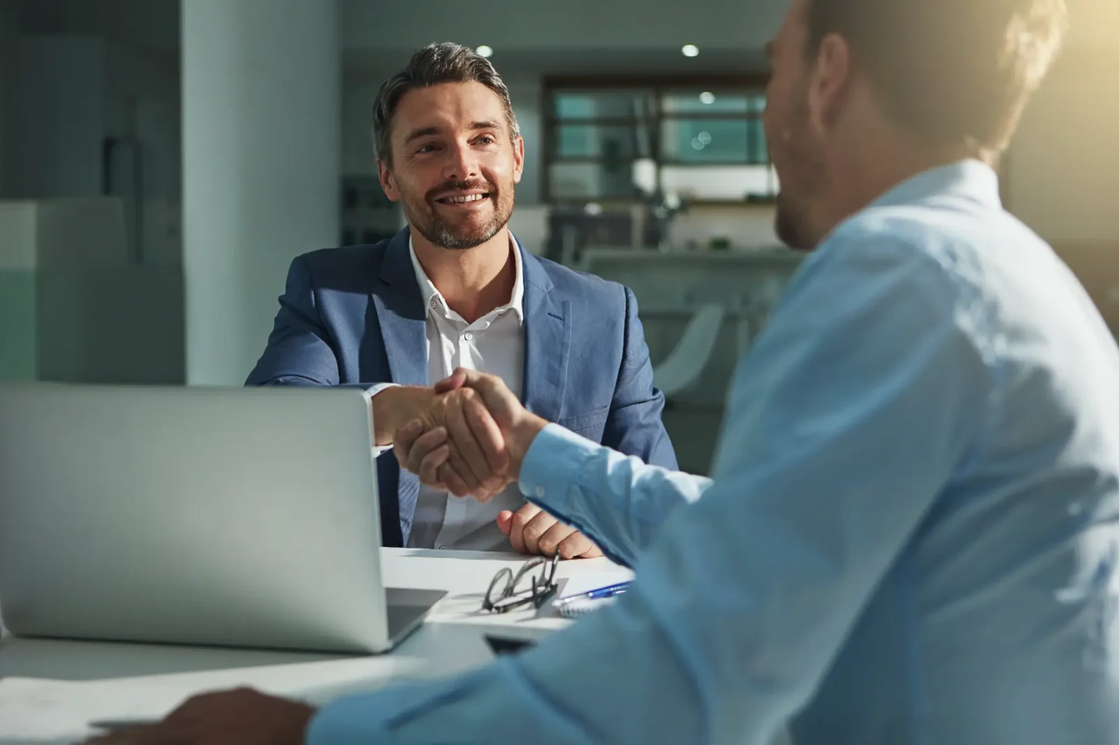 A businessman in a blue suit smiles warmly while shaking hands with a client or partner across a desk with a laptop and documents.