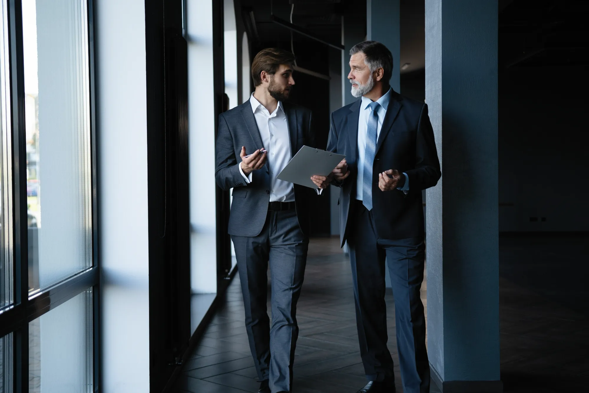 Two businessmen, one younger, one older with a beard, walk down a sunlit office hallway, discussing information on a clipboard.