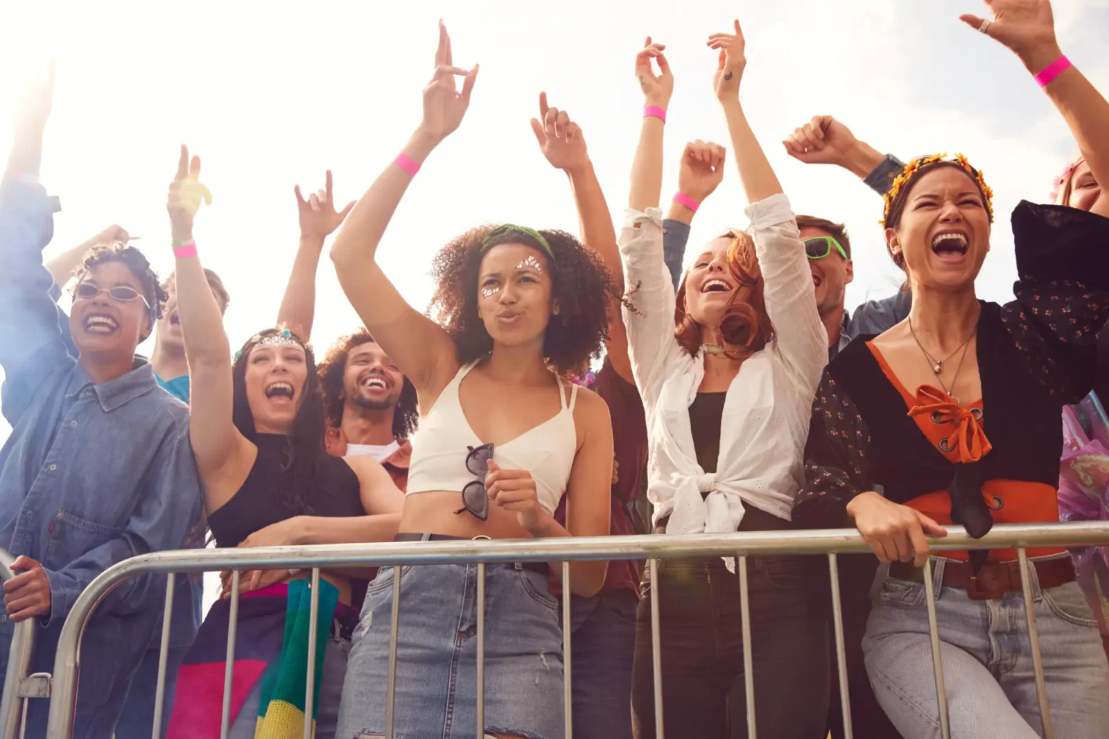 A diverse crowd of excited fans with arms raised stands behind a metal barrier at a sunny outdoor music festival, cheering and laughing.