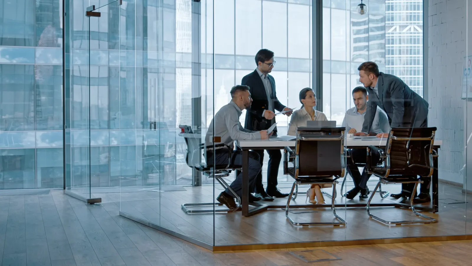 A group of five business professionals meet in a glass conference room overlooking a city. Two standing colleagues present documents to three seated team members.