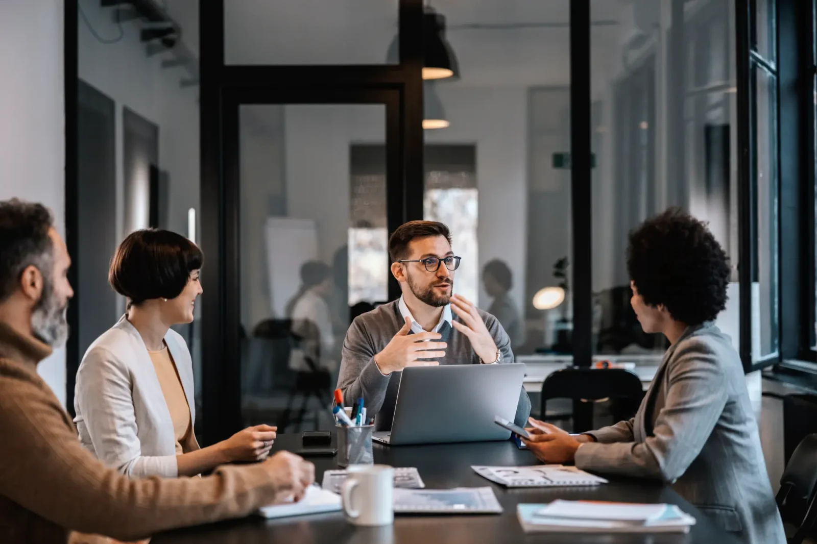 A male professional wearing glasses speaks and gestures during a meeting with three colleagues seated at a conference table. They appear engaged.