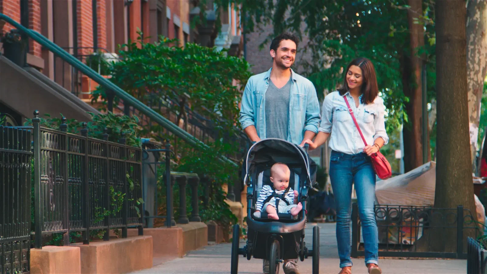 A man and woman, both smiling, walk side-by-side down a city street lined with brownstones. The man pushes a baby strapped into a three-wheeled stroller.