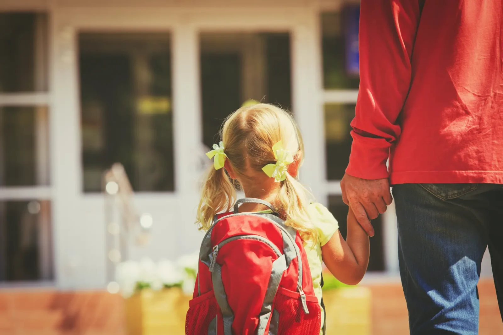 A young girl with a red backpack holds the hand of a parent, walking toward the entrance of a building, likely a school.