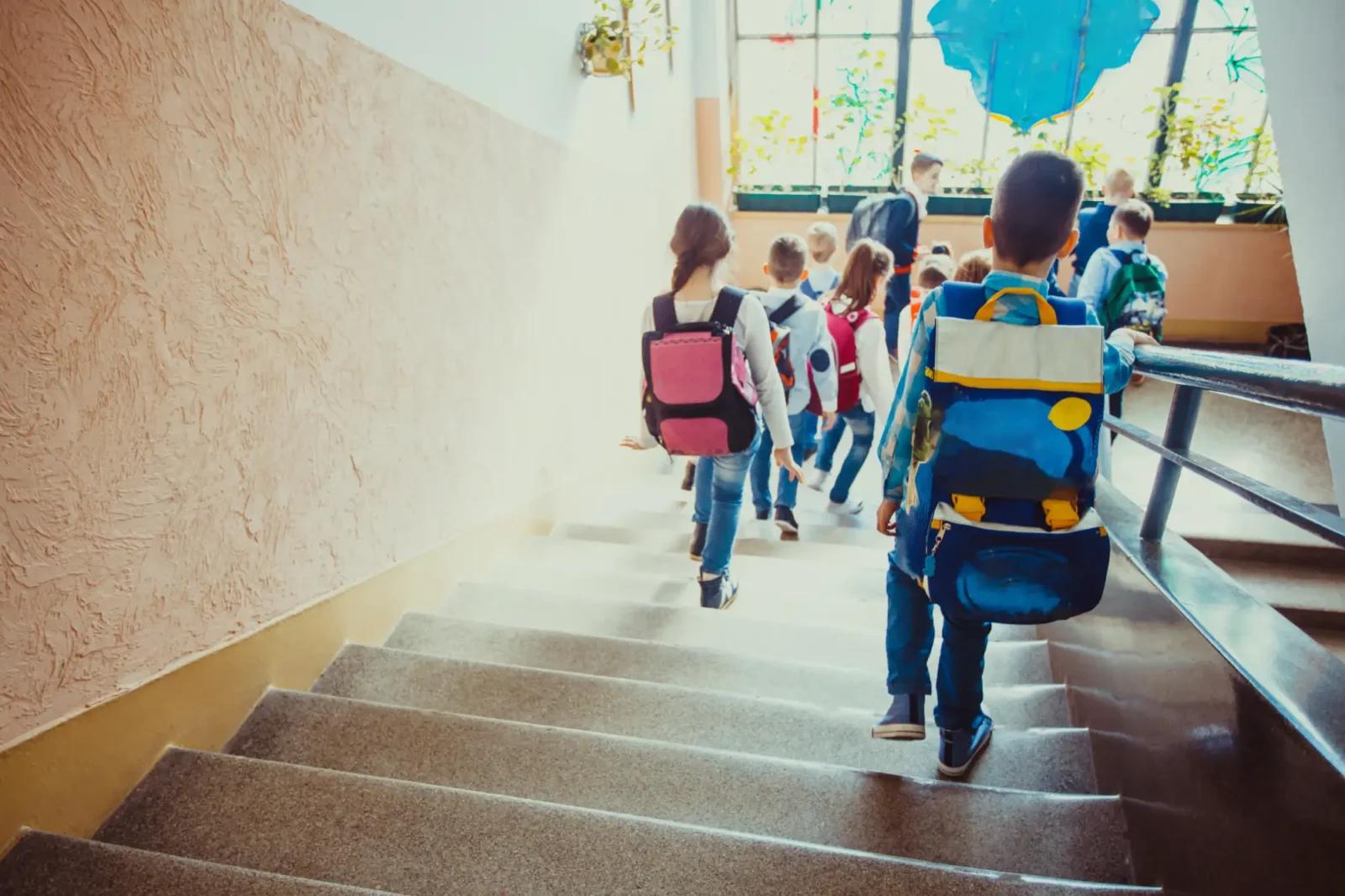 A group of young students with backpacks walks down a granite staircase inside a sunlit hallway, holding onto the metal railing.