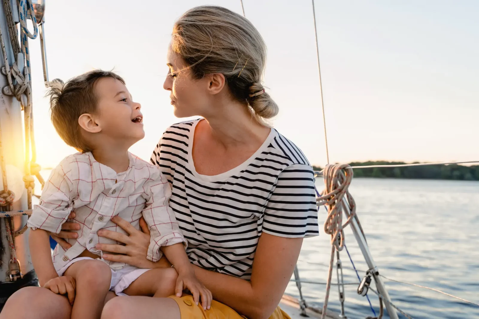 A woman holds her son on a sailboat during sunset. The boy looks up at her with a joyful, open-mouthed smile.