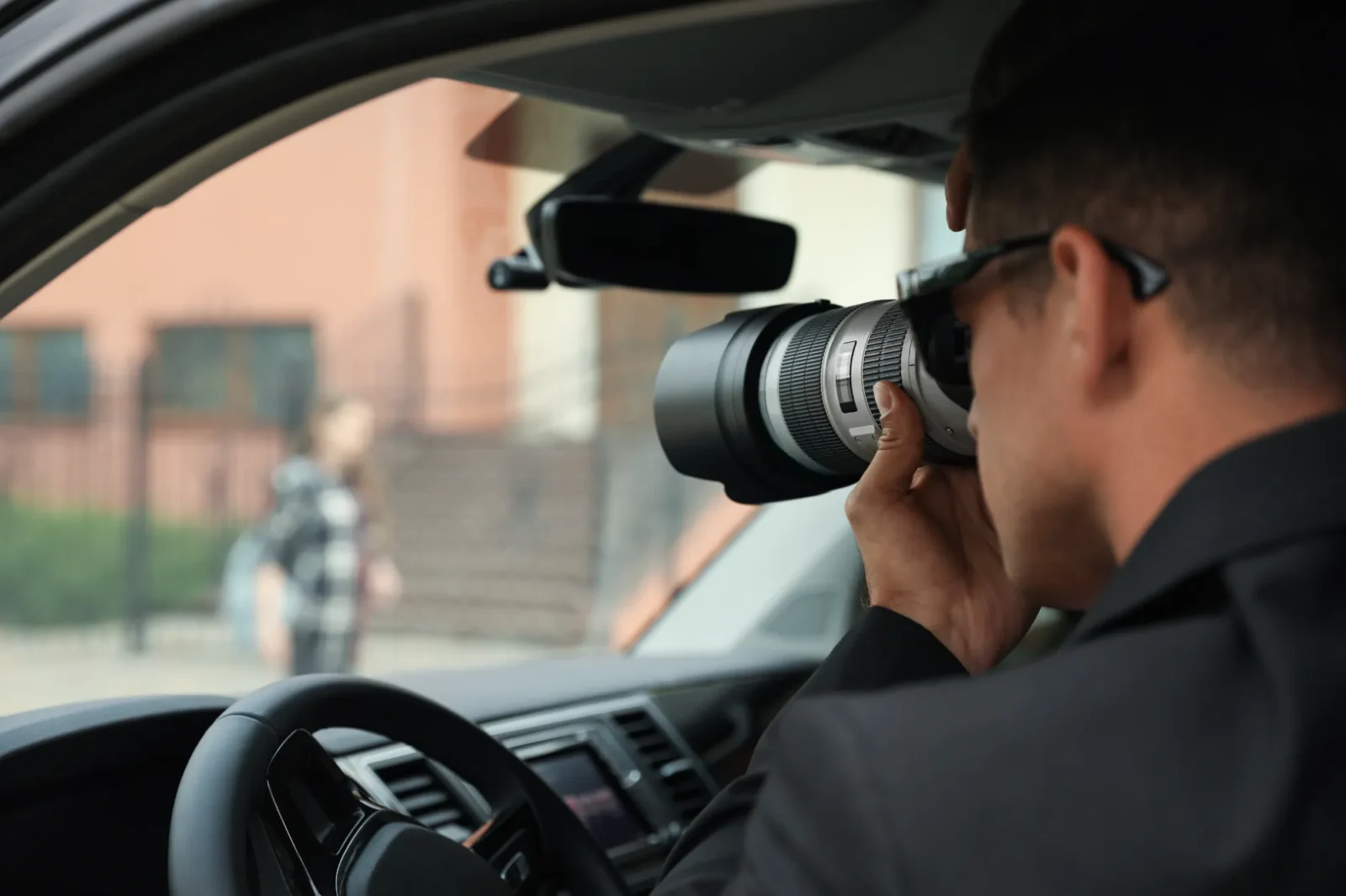 A male private investigator wearing a dark suit and sunglasses uses a large telephoto lens camera while discreetly seated inside a car, observing a blurred subject outside.