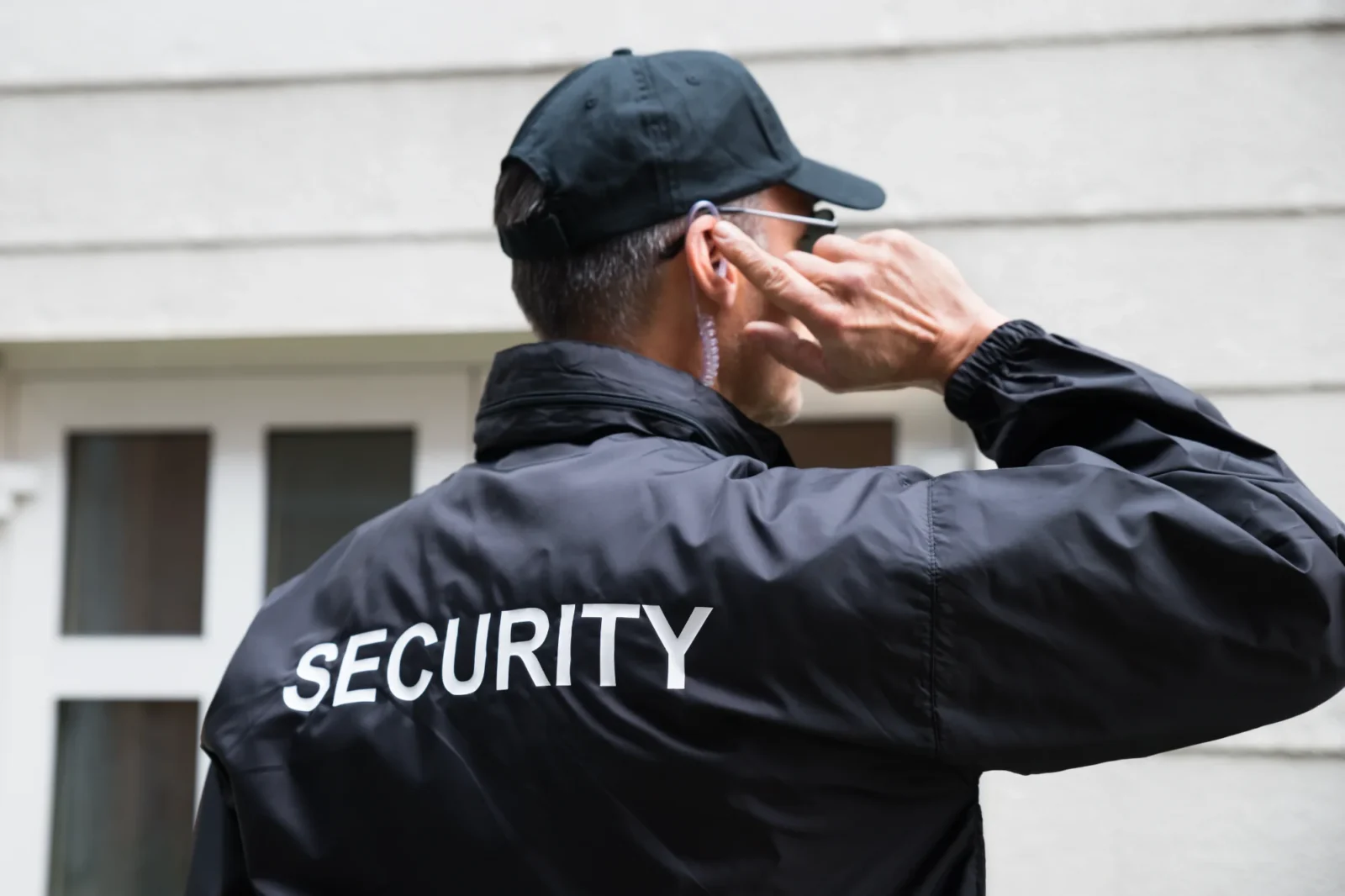 Seen from behind, a security guard in a black jacket and cap touches his earpiece as he communicates outside a white-sided building.