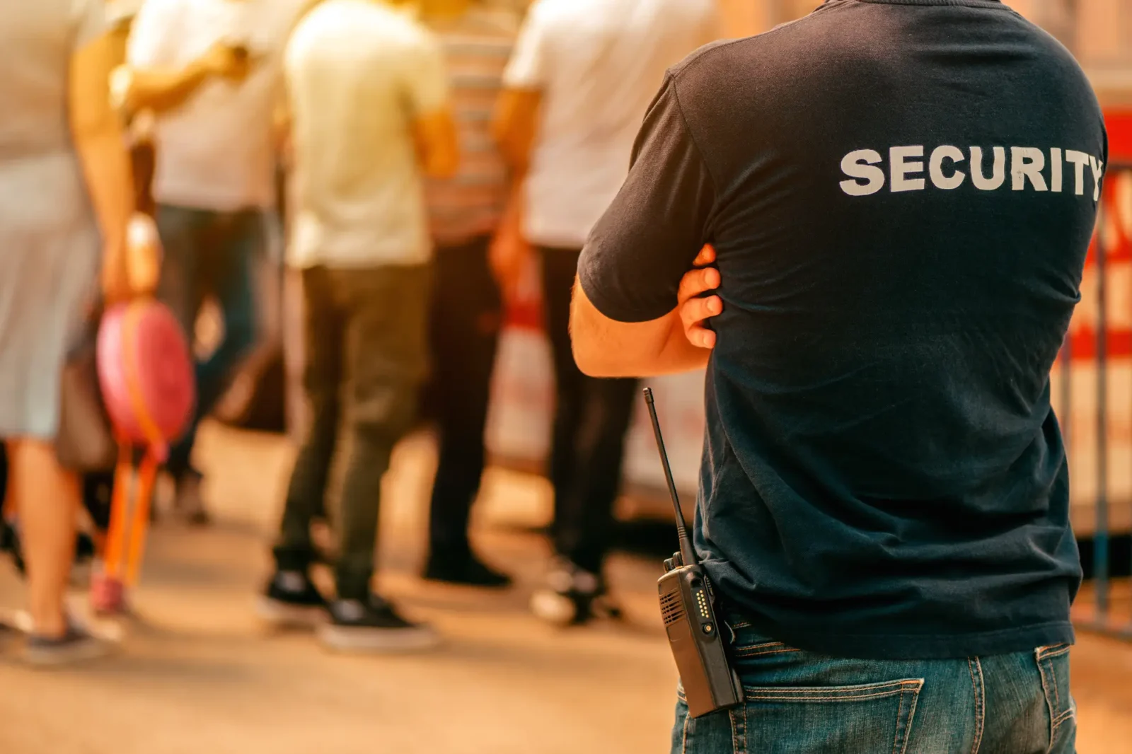 A security guard wears a dark shirt with "SECURITY" on the back and a walkie-talkie clipped to his waist. He stands with his arms crossed, watching a crowd.
