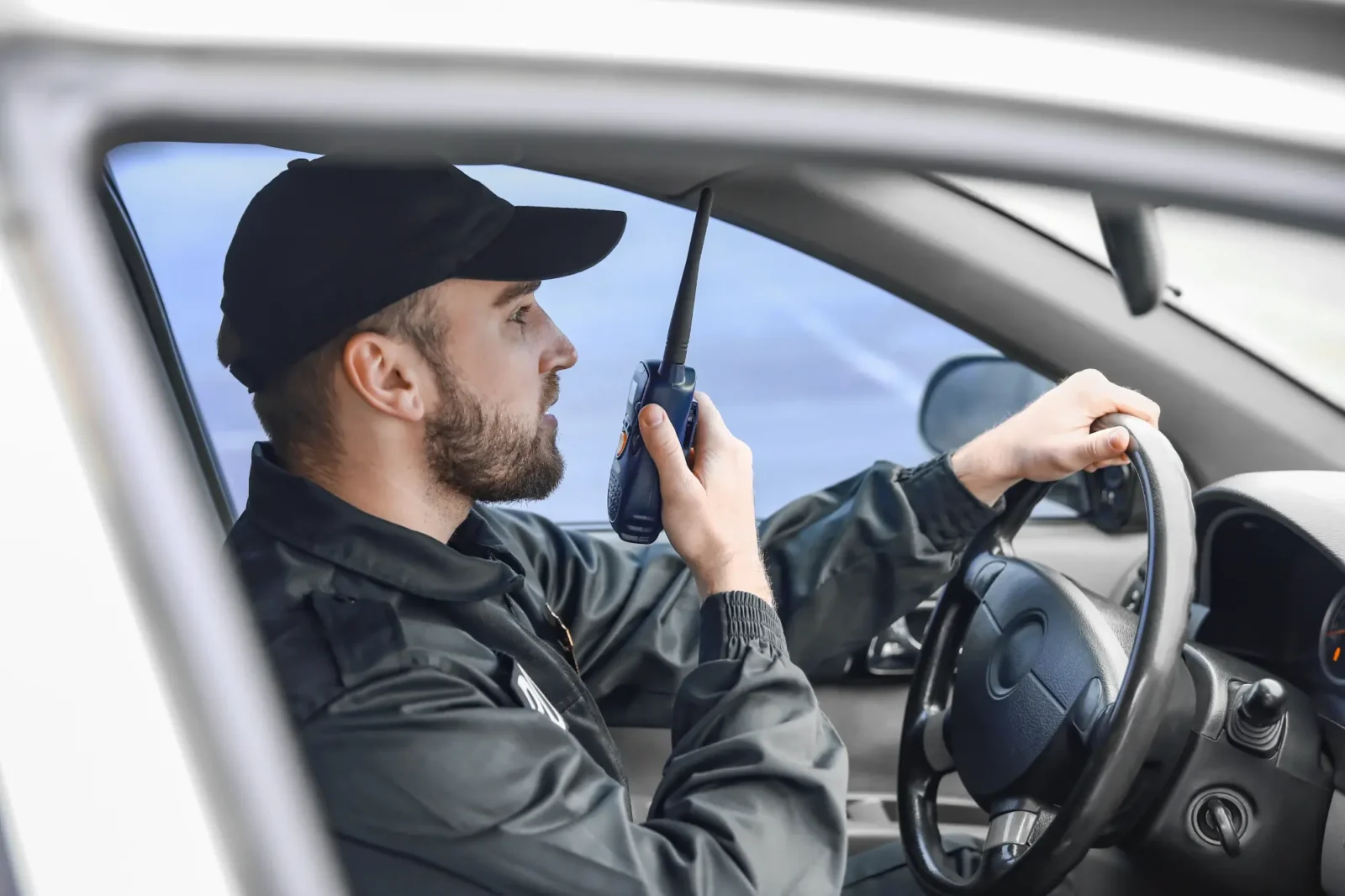 A male security guard in a dark uniform and cap is behind the wheel of a vehicle, speaking into a walkie-talkie with a focused expression.
