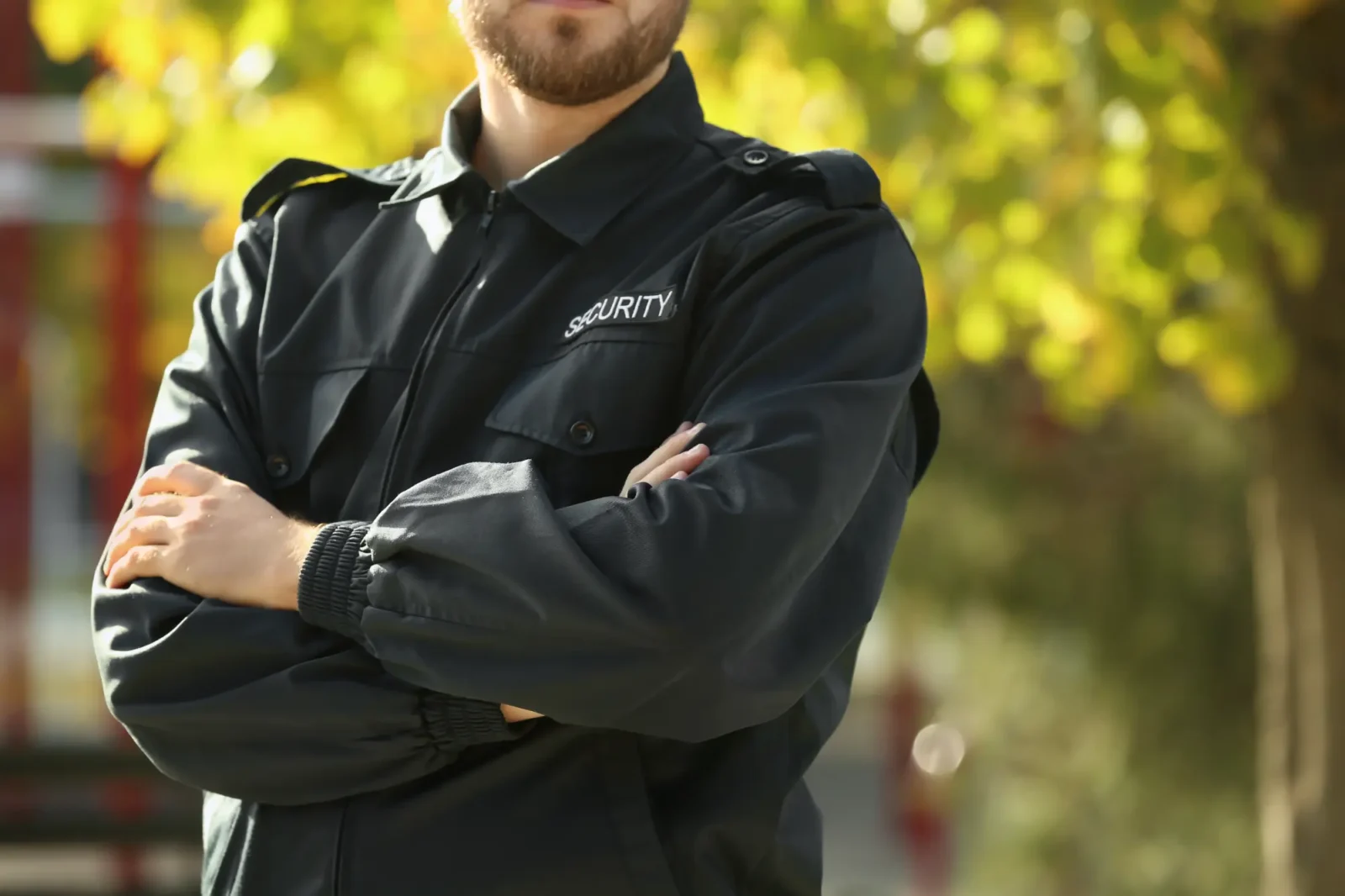 A security guard with a beard, wearing a dark uniform jacket featuring the word "SECURITY" on the chest, stands outdoors with arms crossed.
