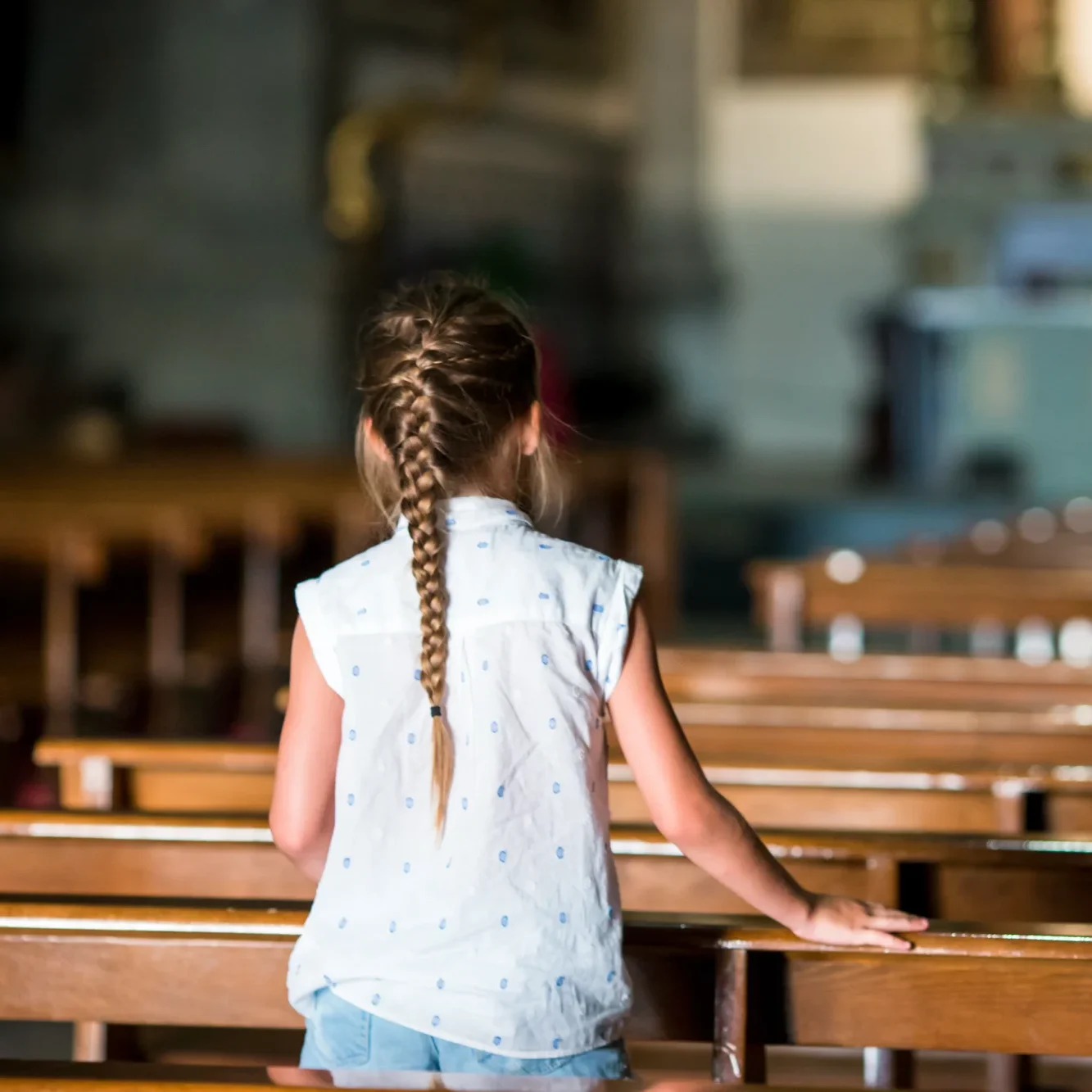 A young girl with a single braid stands in a church aisle, her hand resting on a polished wooden pew. She looks toward the distant altar.