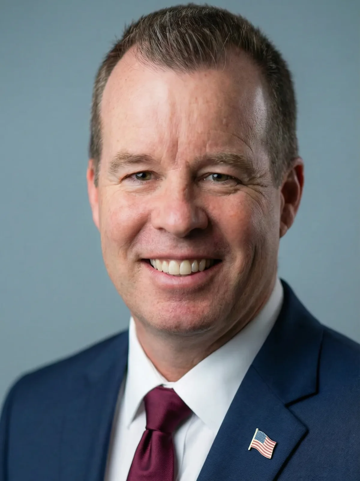 John Ahern, wearing a dark blue suit and tie with an American flag lapel pin, smiles confidently in a professional headshot.